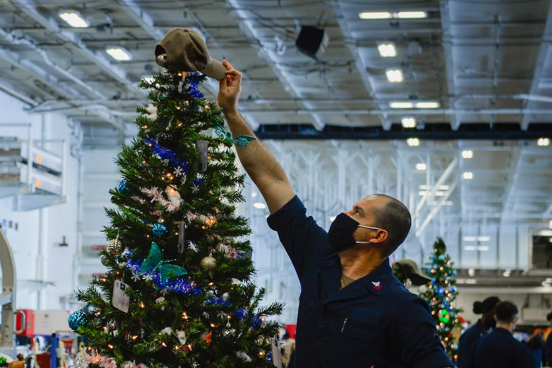 A sailor wearing a mask puts a baseball cap on a Christmas tree.