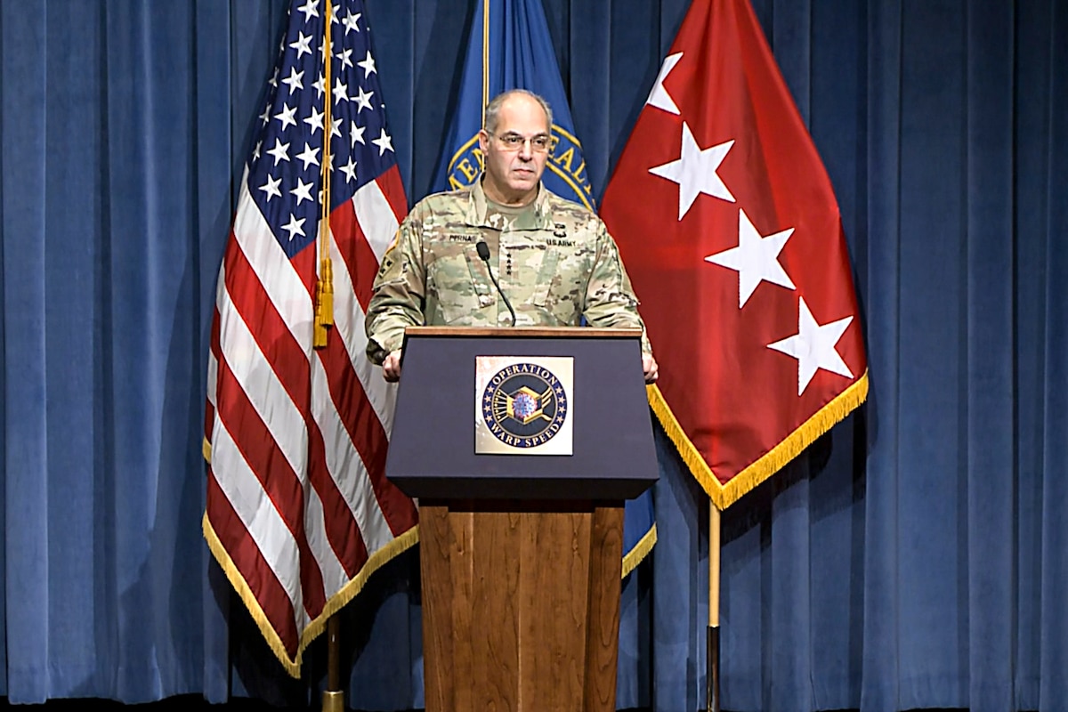 A soldier speaks at a lectern in front of flags.