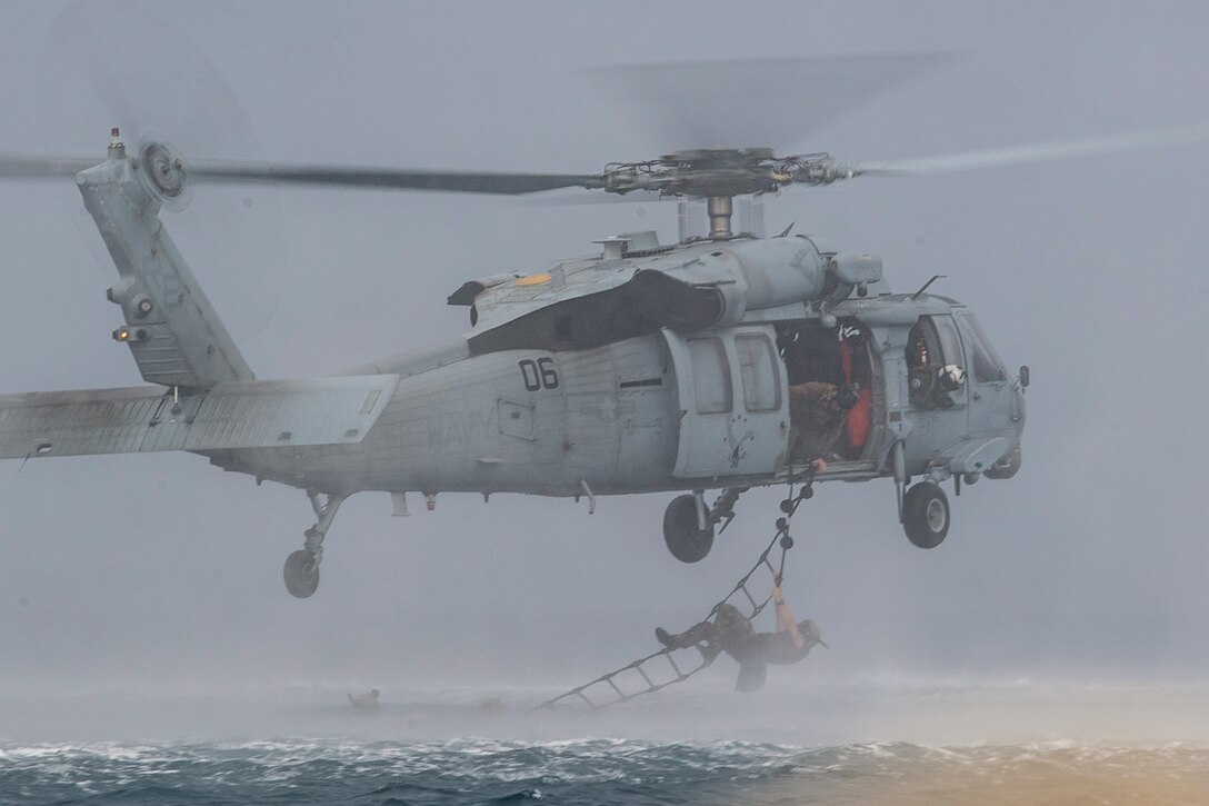 A sailor climbs on a ladder hanging out of an open helicopter hovering over water.