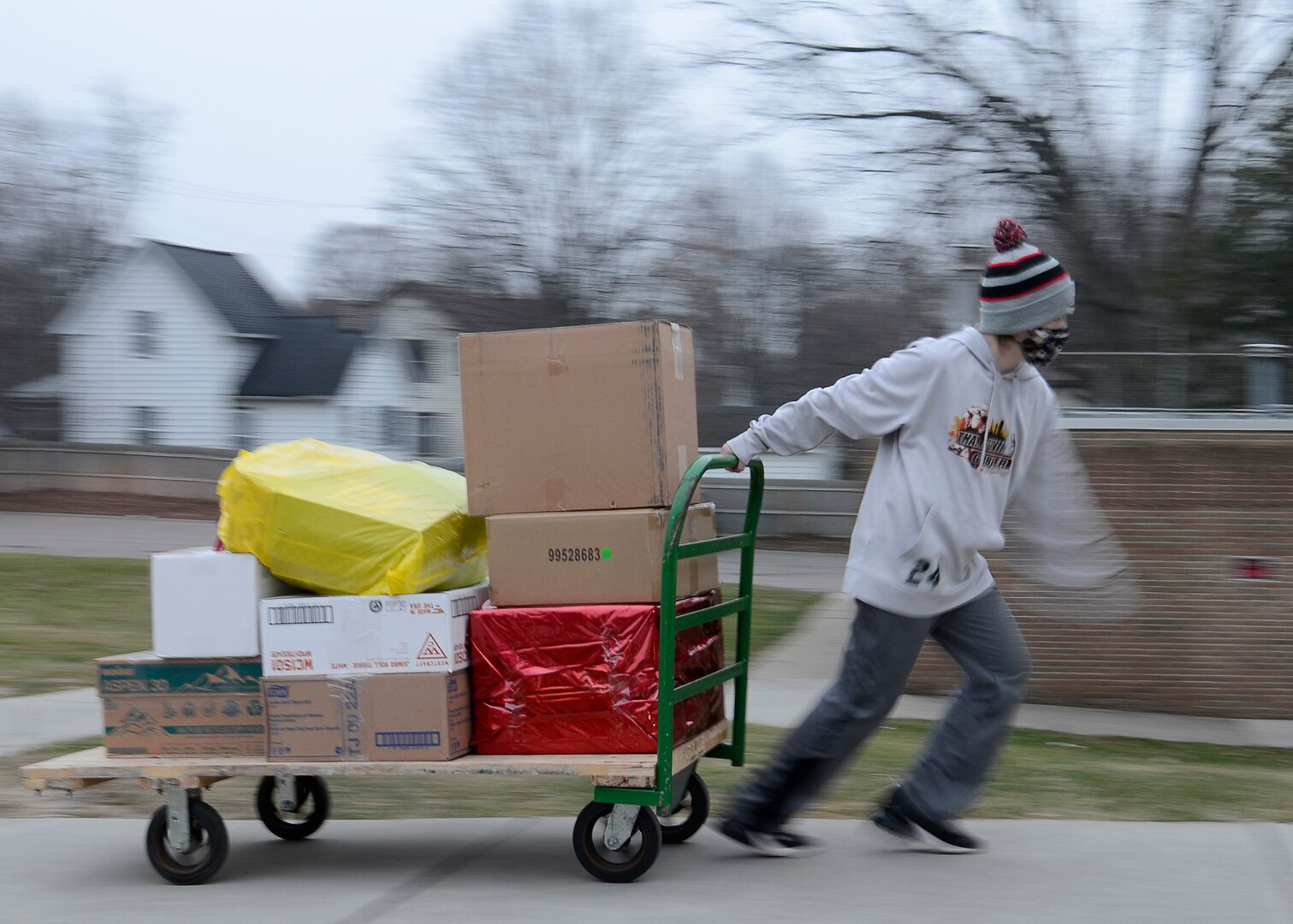 A young boy pulls a hand cart with packages of food and gifts stacked on it towards a door going into a building