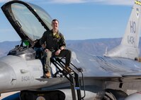 A plot in a green flight suit sits on top of a silver F-16 Viper aircraft in front of a blue sky.