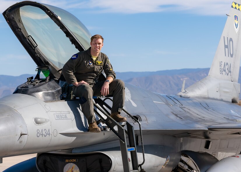 A plot in a green flight suit sits on top of a silver F-16 Viper aircraft in front of a blue sky.