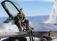 A plot in a green flight suit sits on top of a silver F-16 Viper aircraft in front of a blue sky.