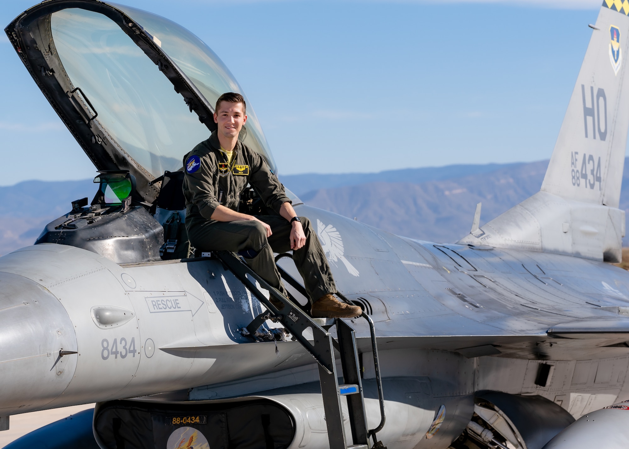 A plot in a green flight suit sits on top of a silver F-16 Viper aircraft in front of a blue sky.