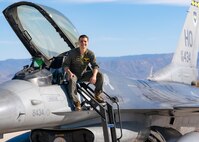 A plot in a green flight suit sits on top of a silver F-16 Viper aircraft in front of a blue sky.
