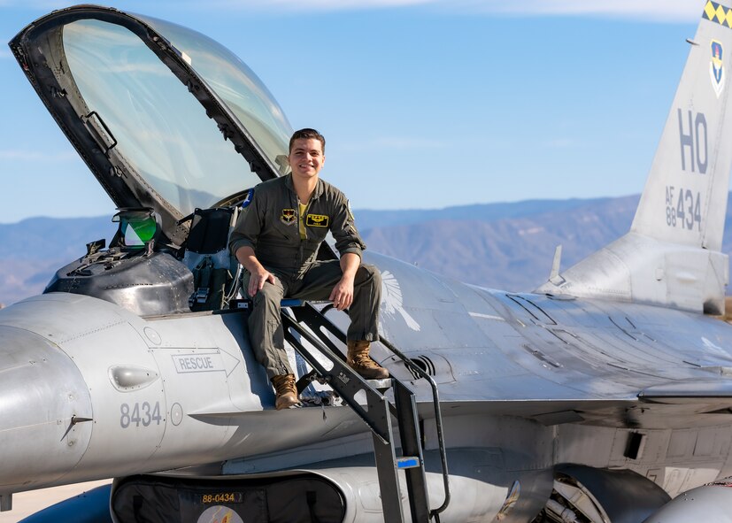 A plot in a green flight suit sits on top of a silver F-16 Viper aircraft in front of a blue sky.