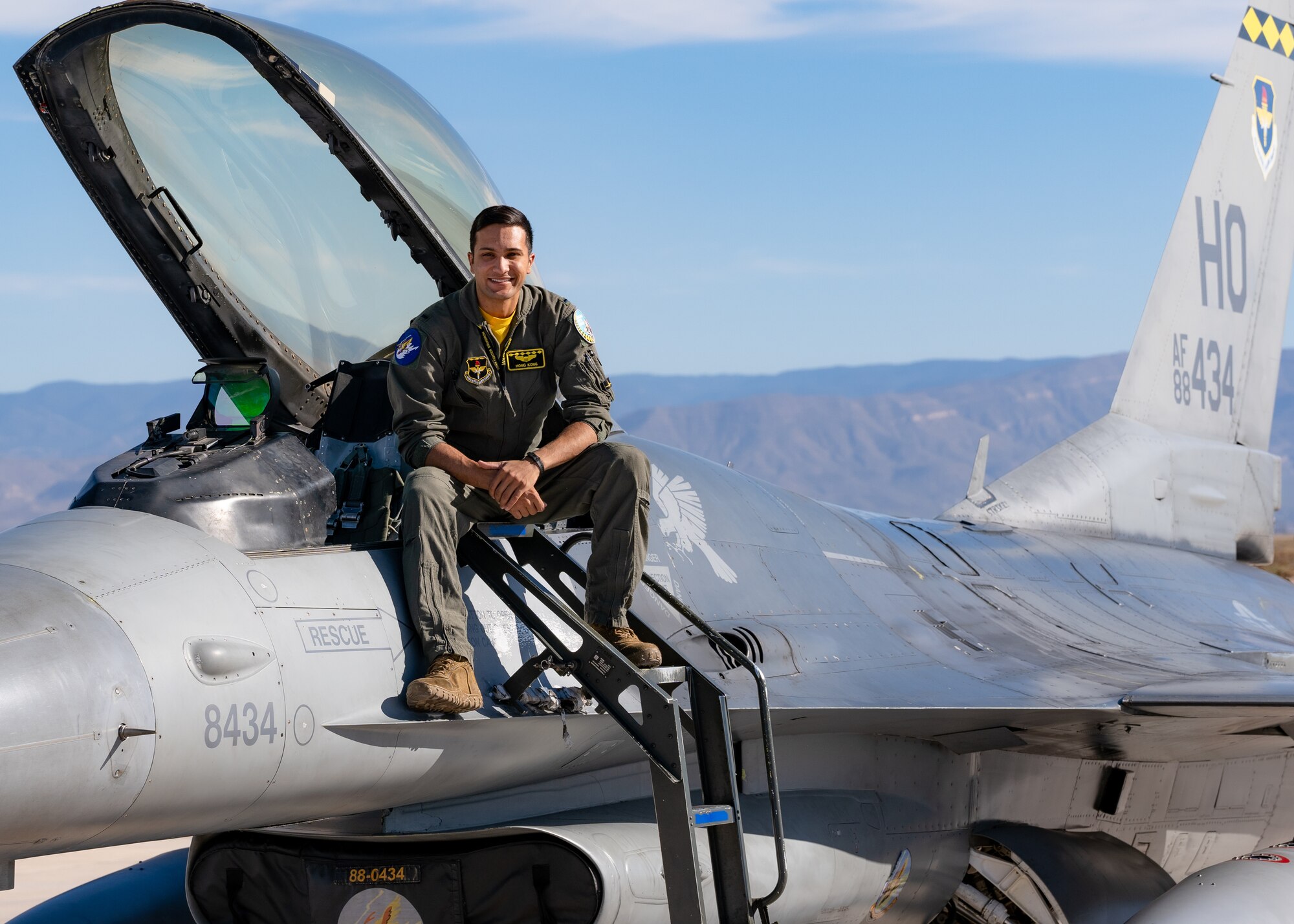 A plot in a green flight suit sits on top of a silver F-16 Viper aircraft in front of a blue sky.
