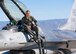 A plot in a green flight suit sits on top of a silver F-16 Viper aircraft in front of a blue sky.