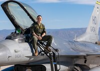 A plot in a green flight suit sits on top of a silver F-16 Viper aircraft in front of a blue sky.