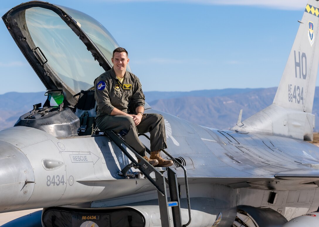 A plot in a green flight suit sits on top of a silver F-16 Viper aircraft in front of a blue sky.