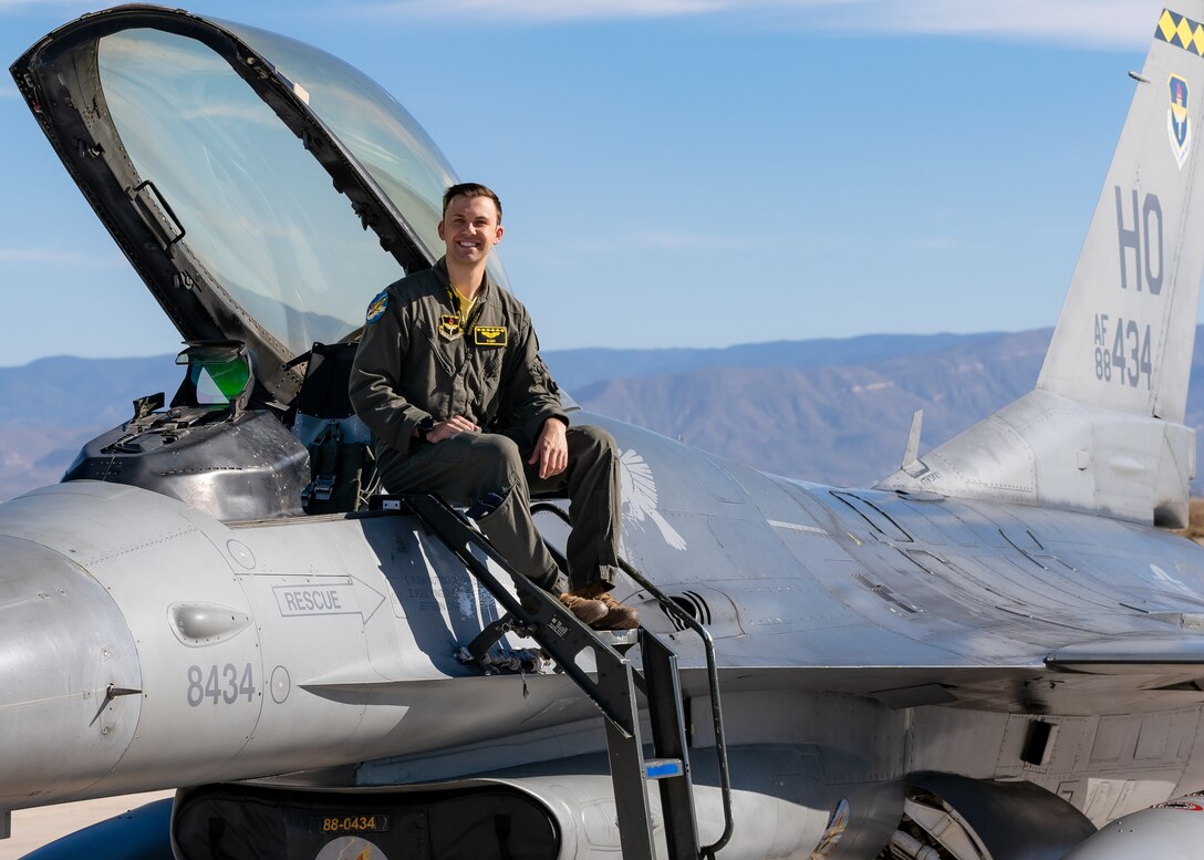A plot in a green flight suit sits on top of a silver F-16 Viper aircraft in front of a blue sky.