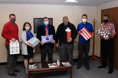Six people stand each wearing a mask and holding a Christmas present in an office setting.