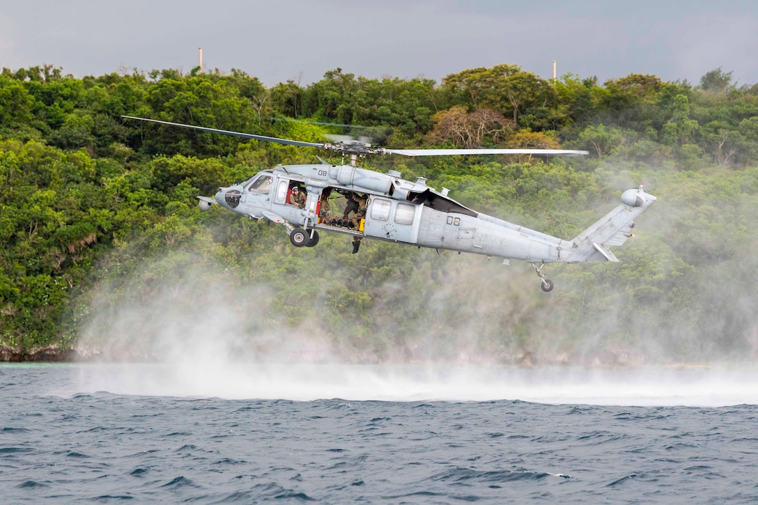 A Navy helicopter hovers over water in front of a tree line.