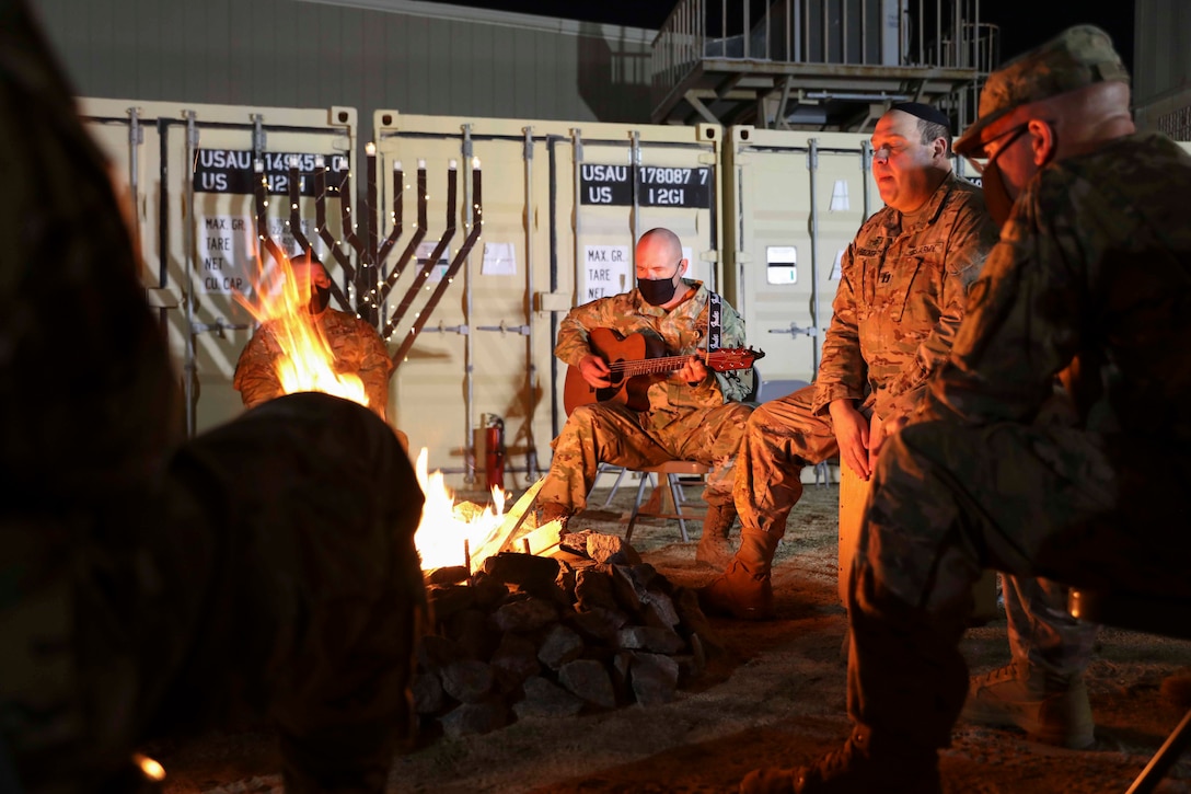 A soldier sits next to a fire while playing a guitar; others sit in a circle around the fire.