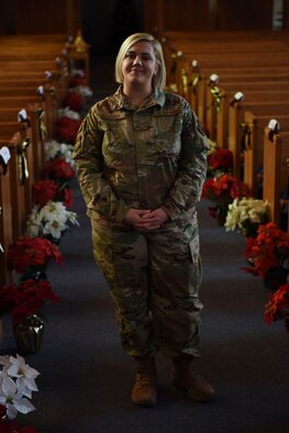 Airman Sara Boucher stands inside of the chapel between the pews, posing for a photo. Flowers line the floor behind her.