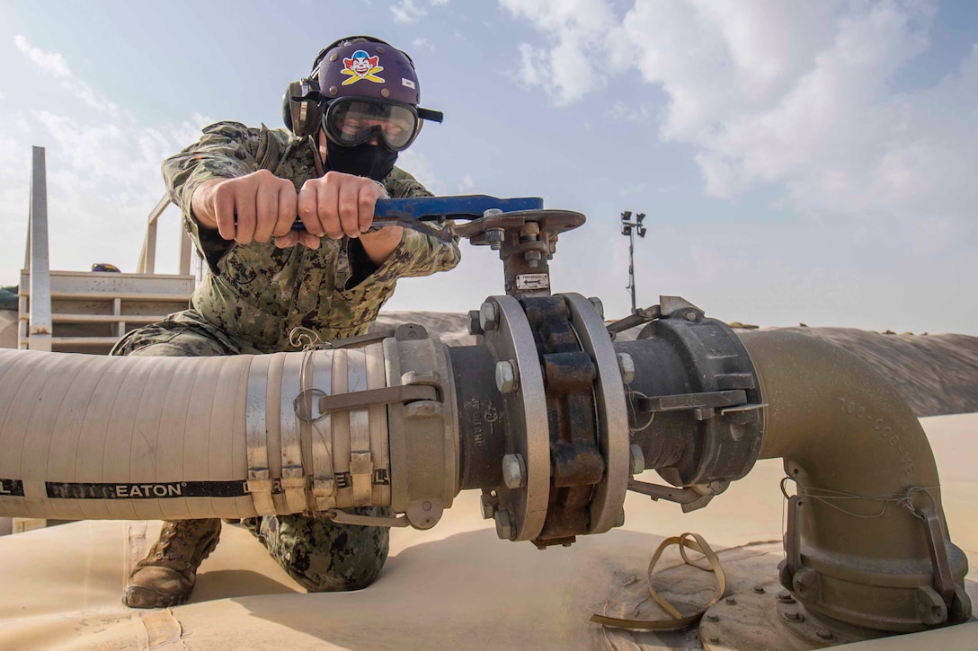 A sailor kneels on the ground while using a wrench to open a valve.