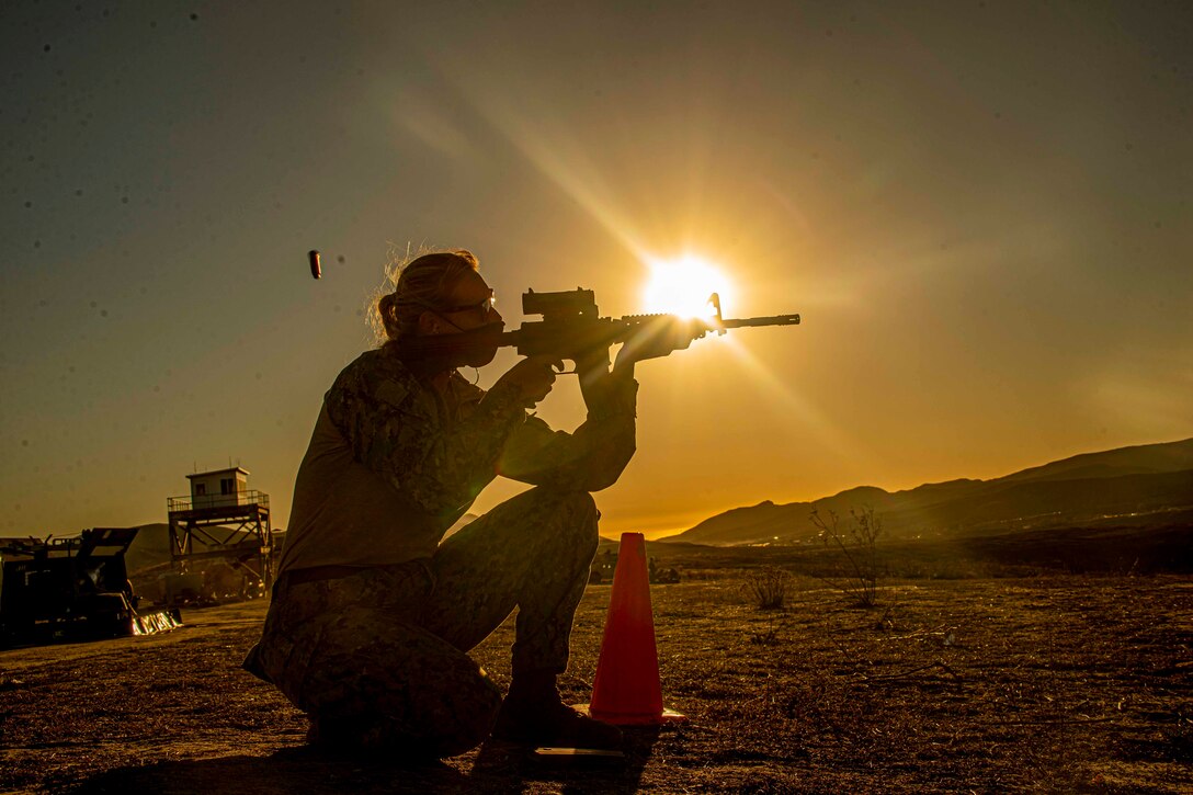 A sailor kneels on the ground while firing a rifle toward a target; the sun shines in the background.