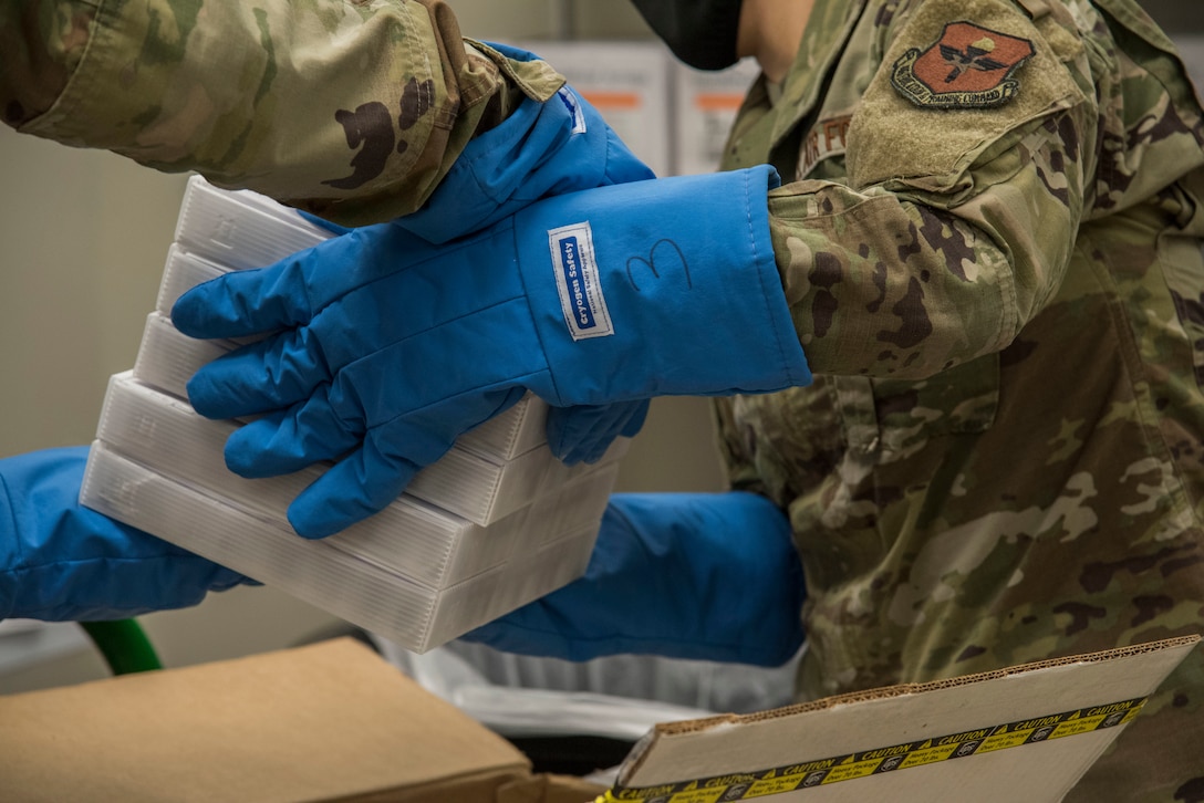Airmen wearing specialty gloves for handling items packed in dry ice pulls out five cases of COVID-19 vaccines from a container.
