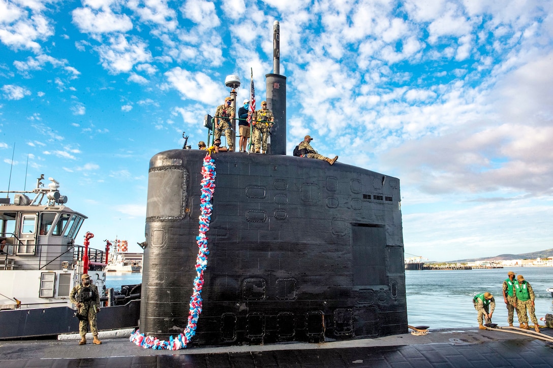 Sailors sit and stand atop a submarine adorned with a lei pulling into a port.
