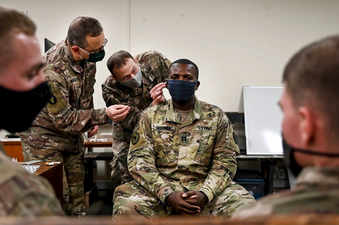 A soldier with an acupuncture needle leans toward another soldier's ear as others watch.