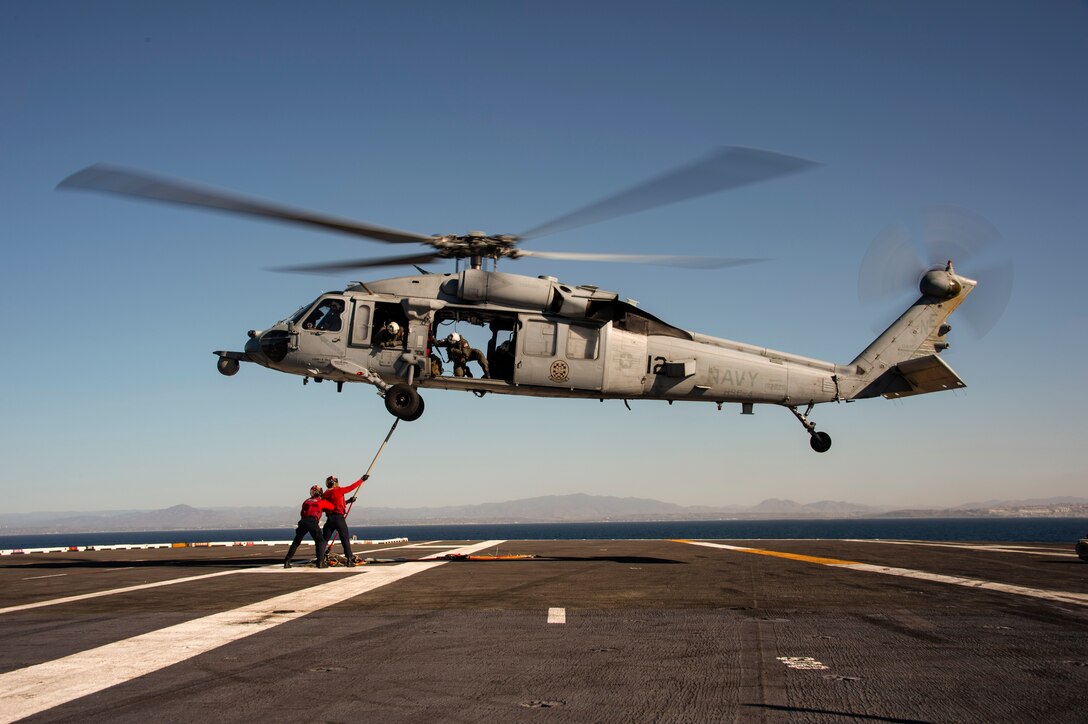 A helicopter hovers over a ship as service members hold a device nearby.