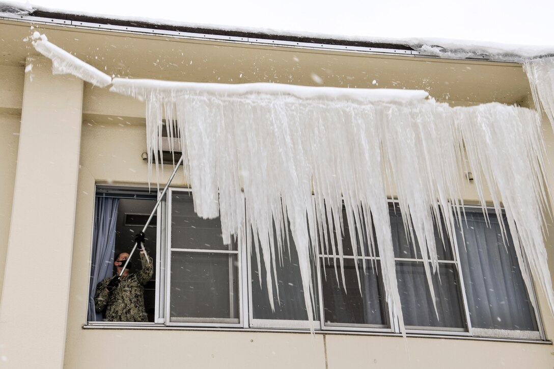 A sailor standing at an open window in a building uses a metal rod to knock down ice from the roof outside.
