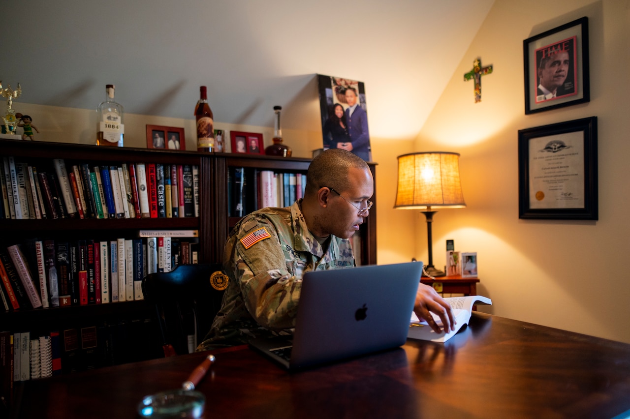 A soldier sits at a desk in front of an open laptop and looks at a book.