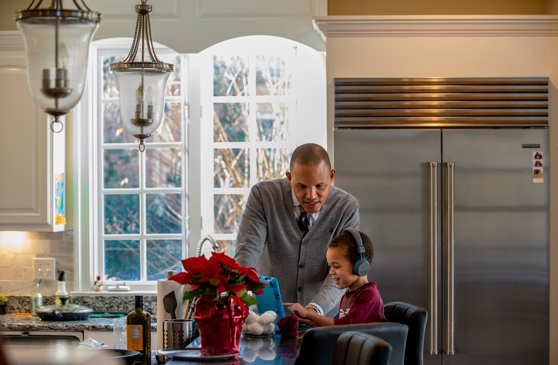 A man watches a boy at a computer wearing headphones sitting at a kitchen counter.