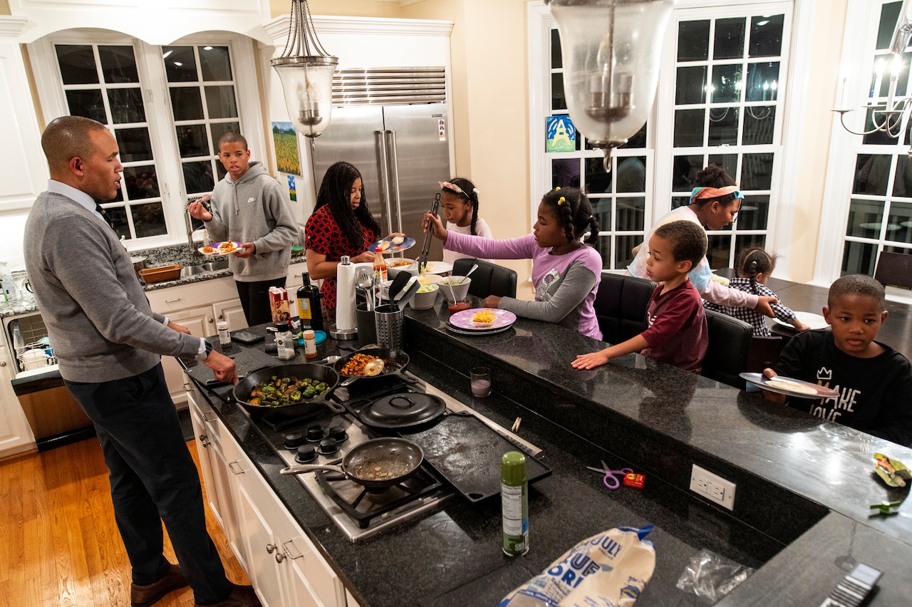 A man stands over a stove as kids hang out in a kitchen.
