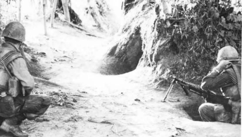 Two men in combat gear crouch down near a shelter’s opening.