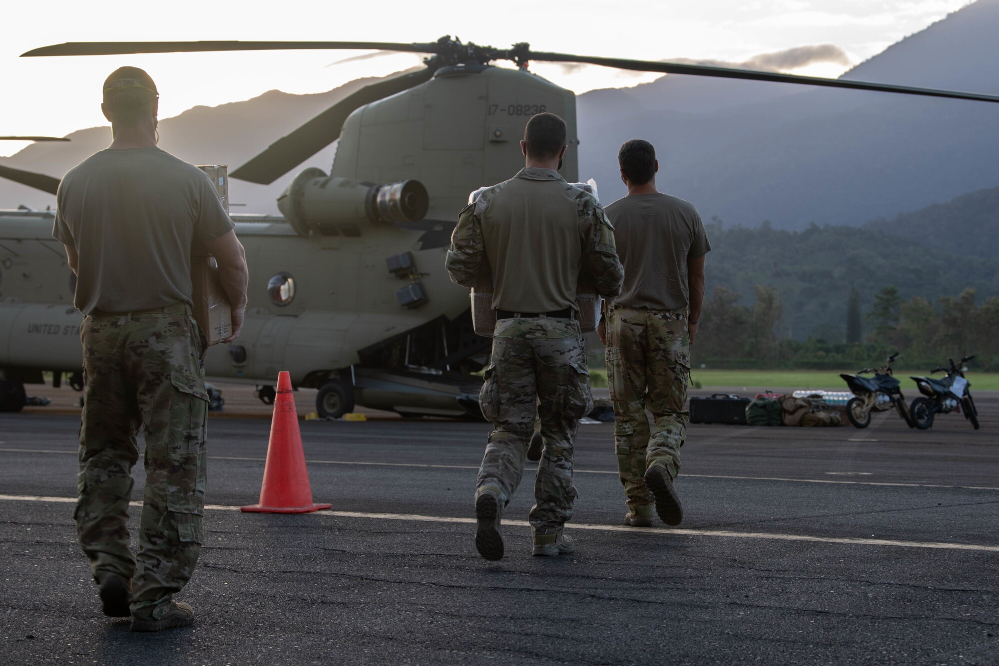 A trio of Special Tactics Operators walk away from us carrying heavy boxes of relief supplies toward the lowered ramp of a large double rotor Chinook helicopter in the background.