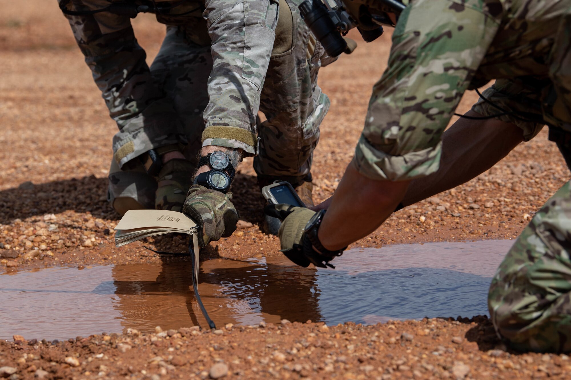 A puddle on a brown gravel runway is shown with the hands of two Special Tactics operators shown on the right measuring the water's depth.