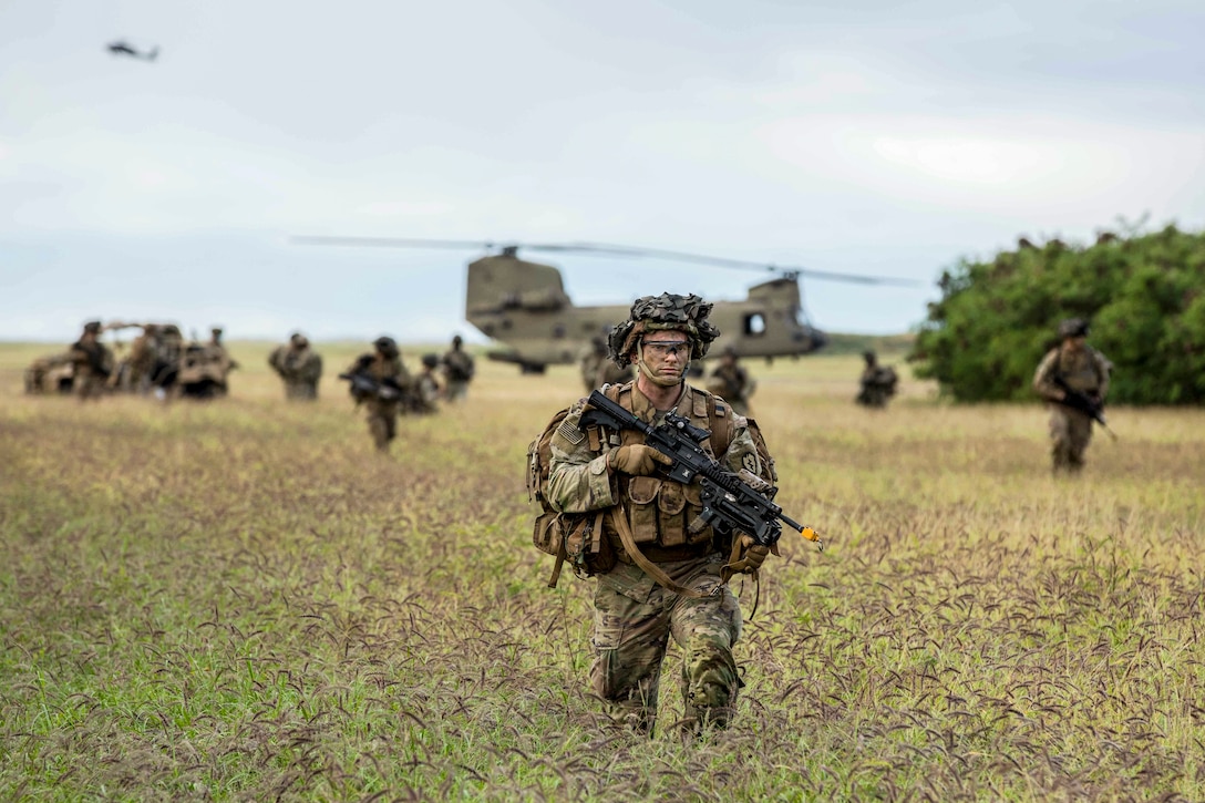 A soldier walks with a weapon through a field as others walk in the background near a parked helicopter.