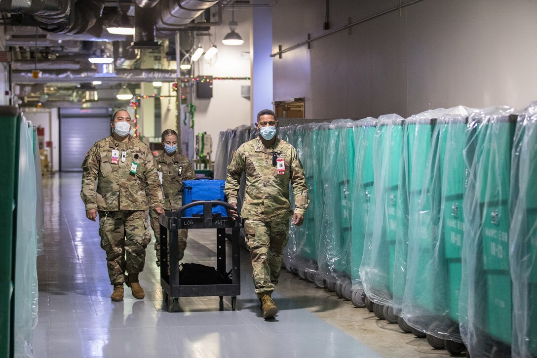 Three soldiers wearing face masks push a cart with a container holding COVID-19 vaccines down a hallway.
