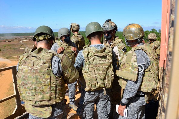 We look over the shoulders of a group of three Colombian special operators  wearing tan protective vests and green helmets. They are listening to a Special Tactics operator facing us from the other side of this trio. The group is standing on a ledge looking over a valley with green forest stretching off into the distance. A Special Tactics operator with radios on his vest stands beyond this small group.