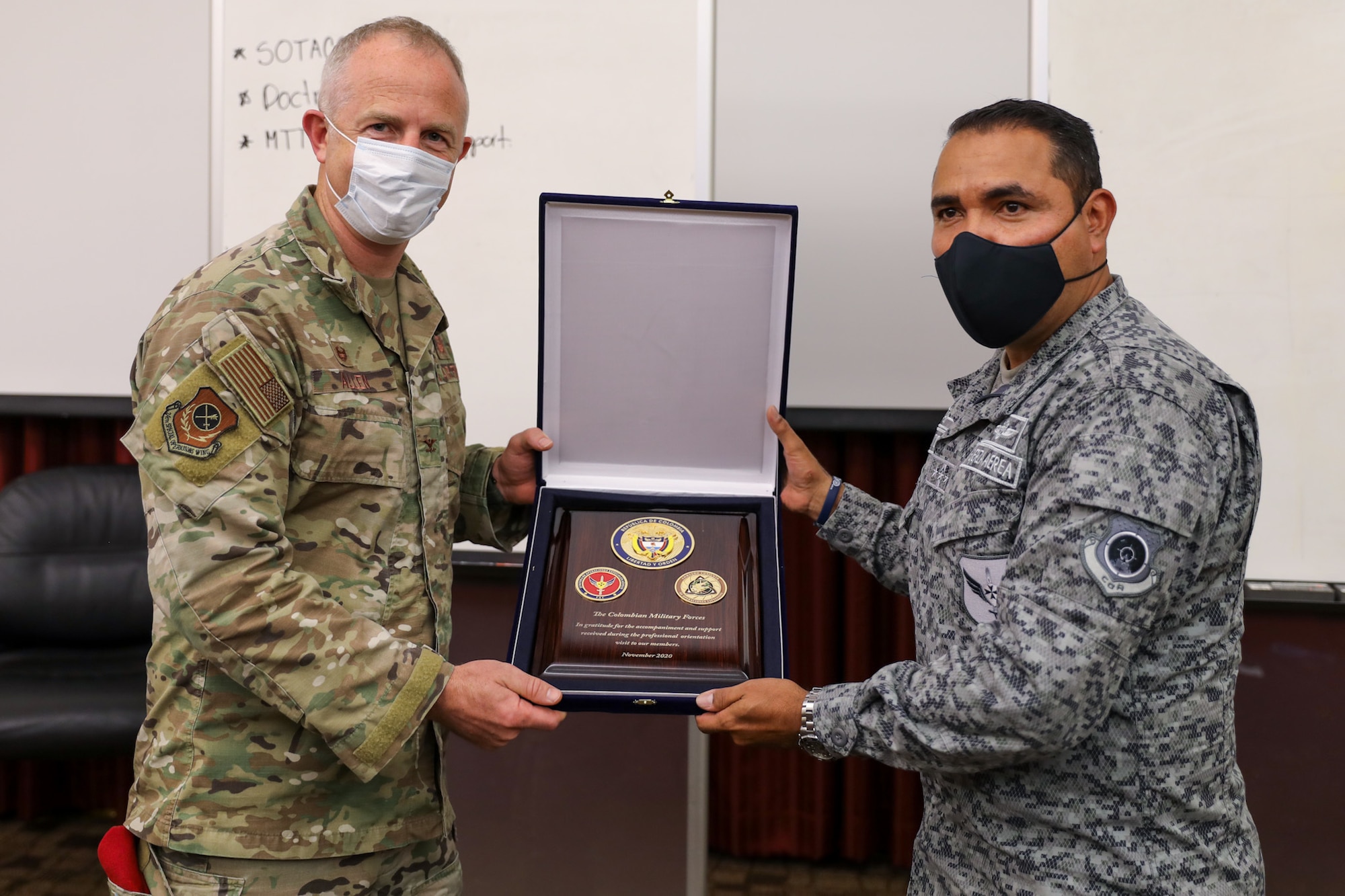 An Officer of the Special Tactics stands on the left side of the picture, and an Officer of the Colombian Air Force stands on the right side of the picture, both are masked for COVID-19. Between them they hold up a large felt-covered box containing a wooden plaque being presented by the Colombian military to the Special Tactics. There are three unit insignias on the plaque and words in English thanking the Special Tactics.