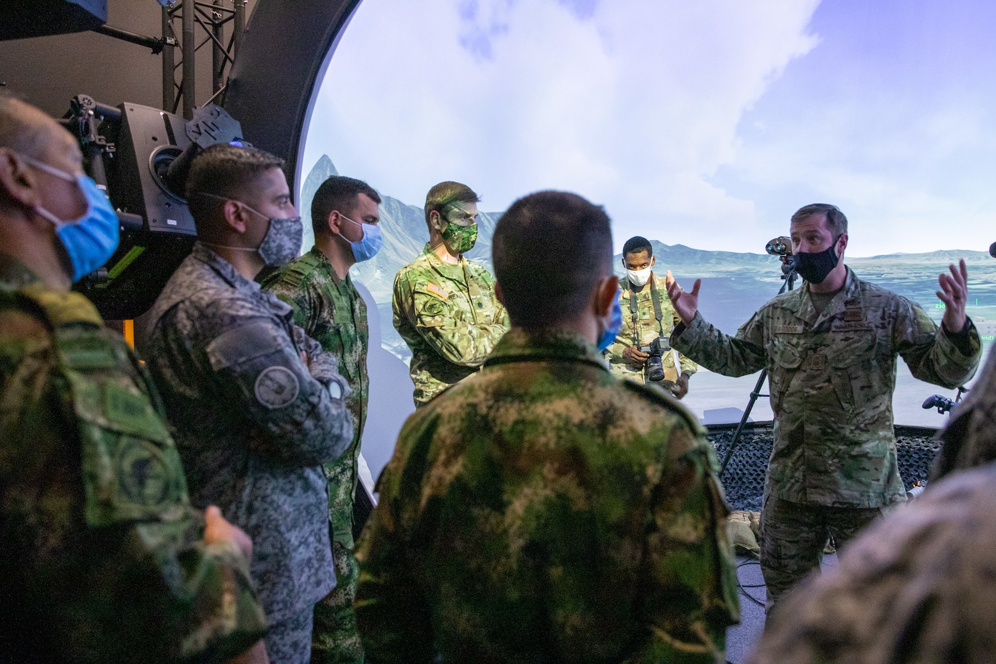 A Special Tactics operator in a black COVID-19 mask faces the camera in front of a video screen. The operator is addressing members of the US and Columbian military in camouflaged uniforms and masks.