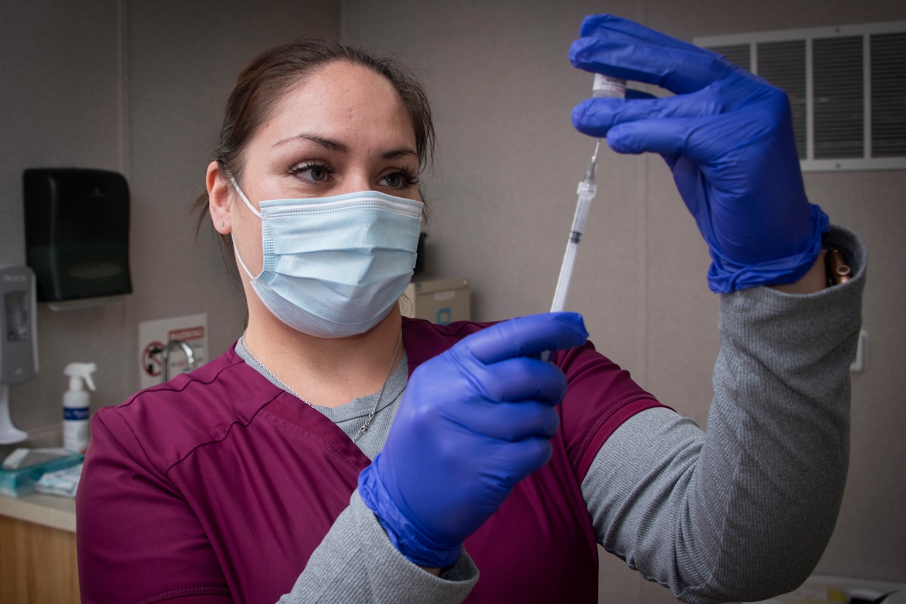 A woman wearing a mask uses a syringe to draw fluid from a small bottle she holds in her gloved hands.