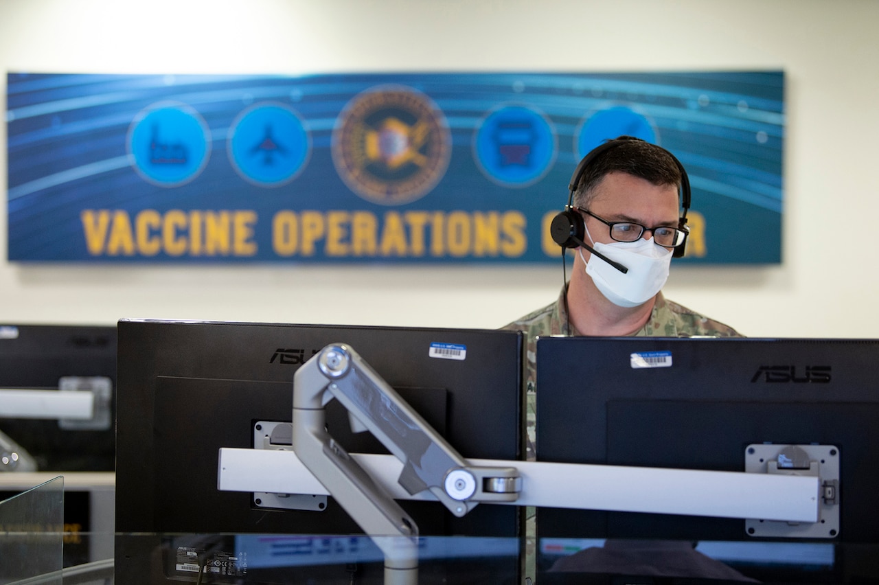 A man wearing a face mask and headset looks at two computer monitors.