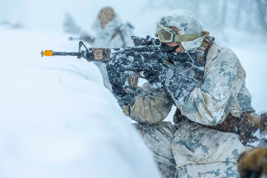 A Marine looks through the scope of a weapon while kneeling behind a mound of snow.