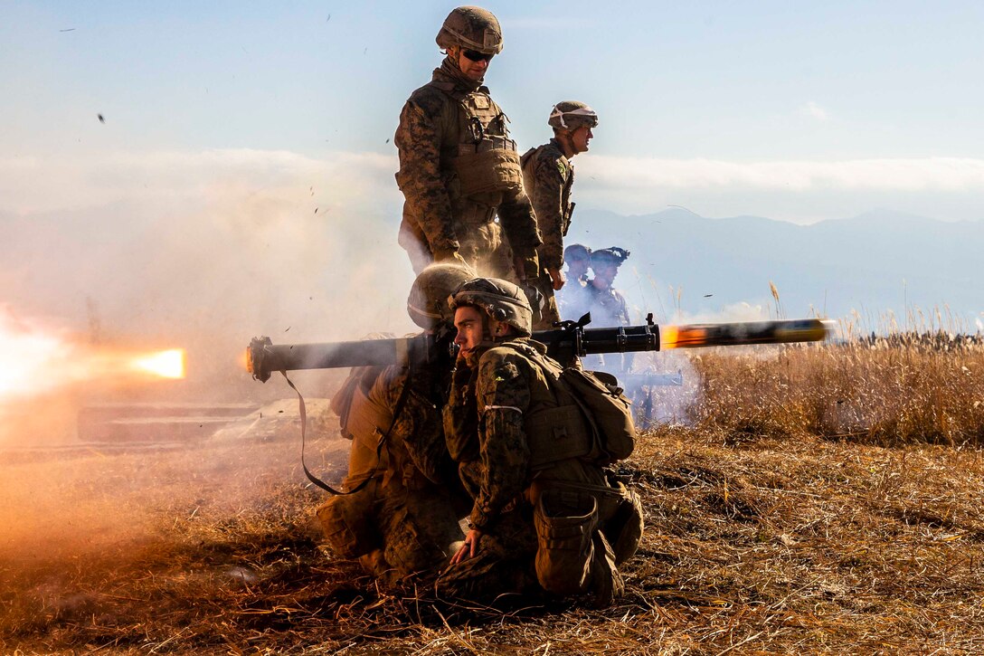 Two Marines kneel on the ground and fire a weapon from their shoulders; others stand behind.