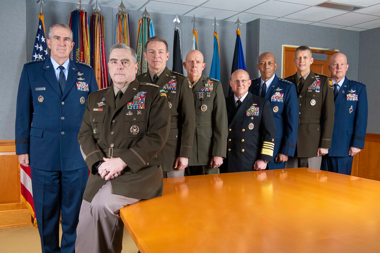 Eight men wearing military uniforms gather around a table; a number of flags are in the background.