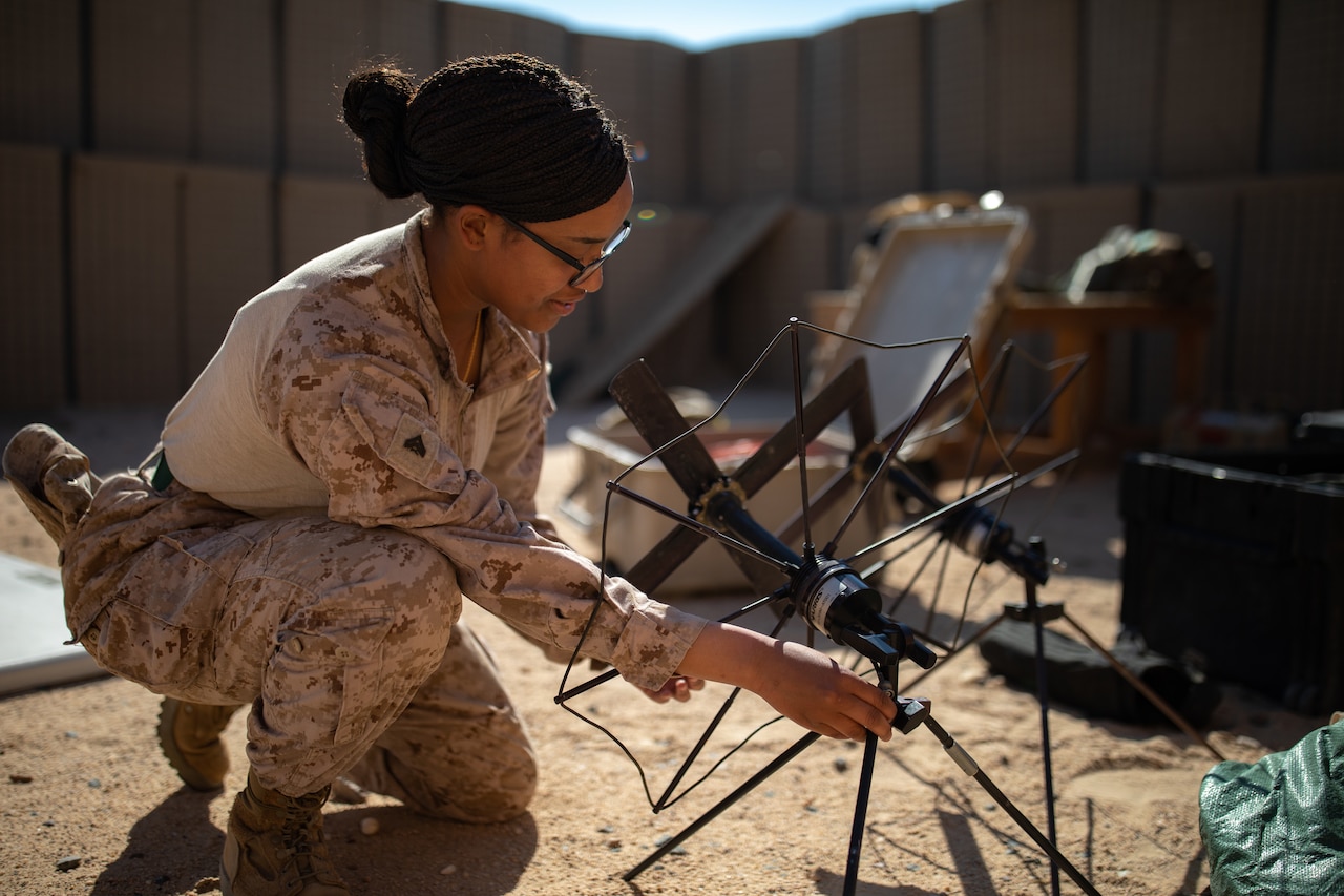 A woman wearing a military uniform crouches on the ground as she sets up a satellite; an equipment case is open behind her.