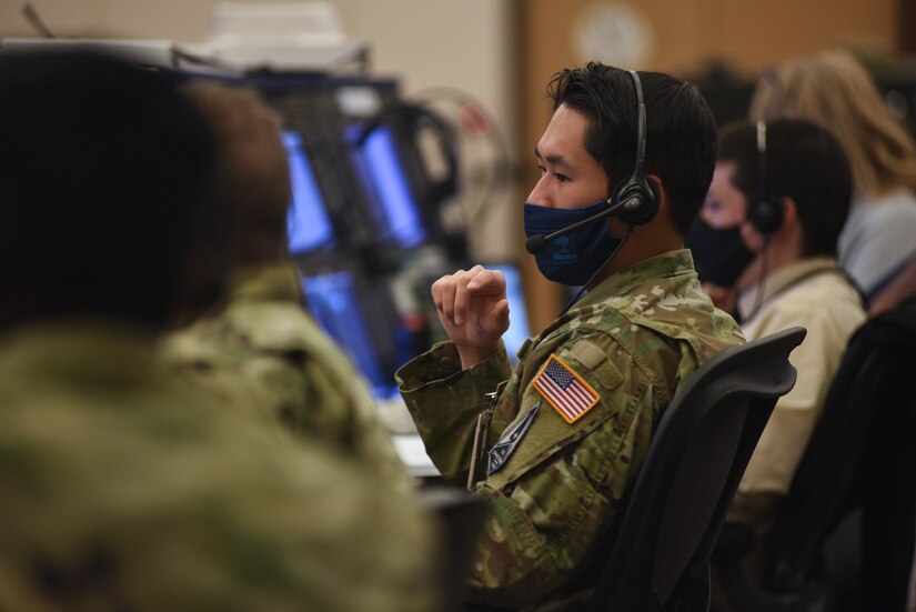 A row of people wearing headsets and face masks sit at desks and look at computer screens.