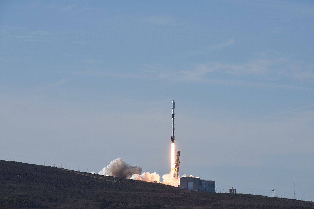 A rocket launches into a bright blue sky as smoke billows beneath it.