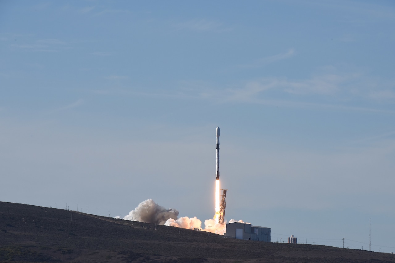 A rocket launches into a bright blue sky as smoke billows beneath it.