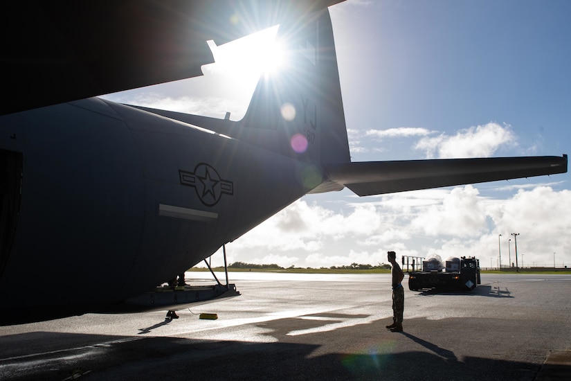 An airman stands on an airstrip; a large plane, ready to accept cargo, is on the left.