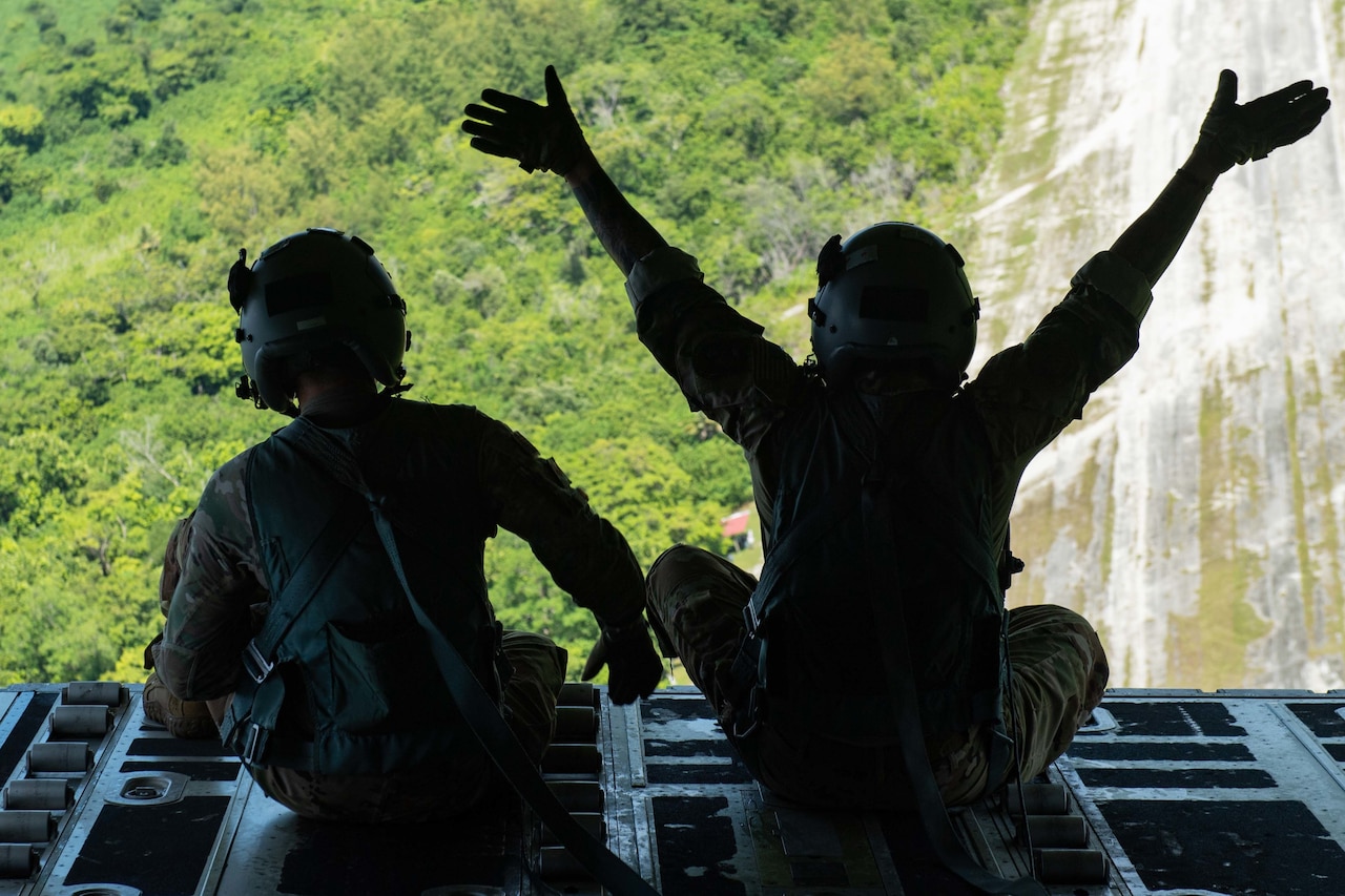 Two people are photographed waving from the open back of a cargo plane.