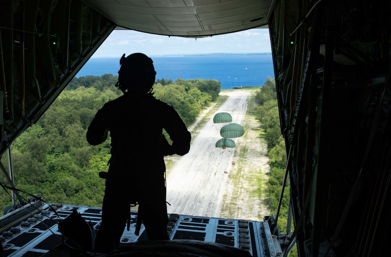 A person wearing a helmet watches bundles airdropped from a cargo plane as they parachute to the ground.