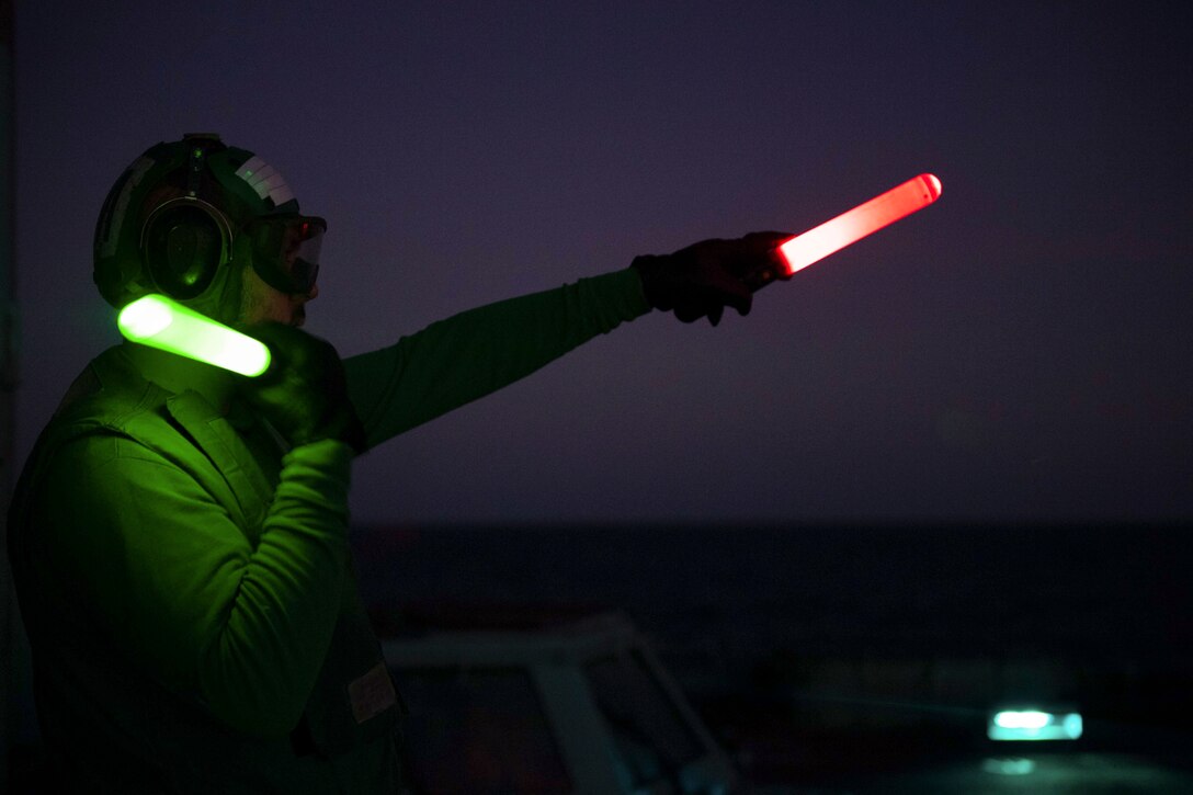 A sailor signals a helicopter illuminated by green and red light.